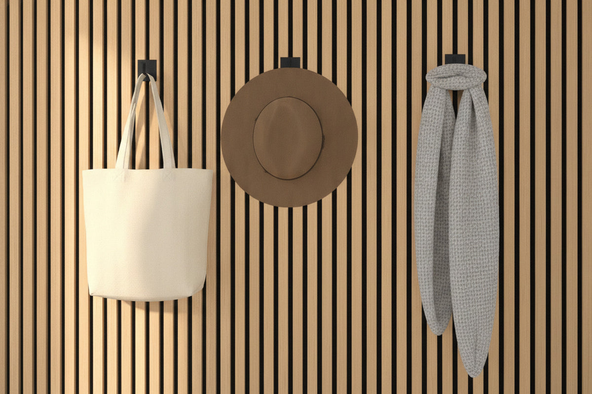 Three identical matte-black steel wall hooks on an oak slat wall holding a canvas tote, a felt hat and a knit scarf
